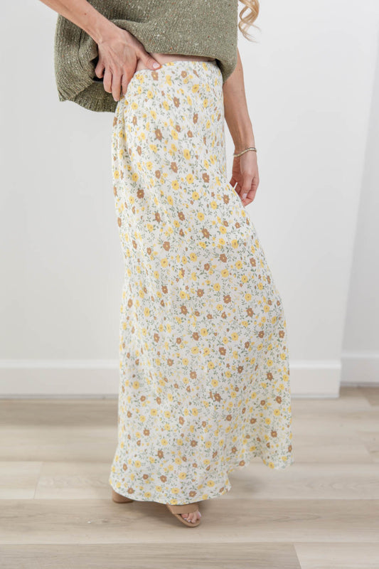 A woman wearing a long, flowing maxi skirt with a small yellow, brown, and blue floral print on a cream background, paired with a textured olive green top. She is standing barefoot on a light wood floor against a white wall.