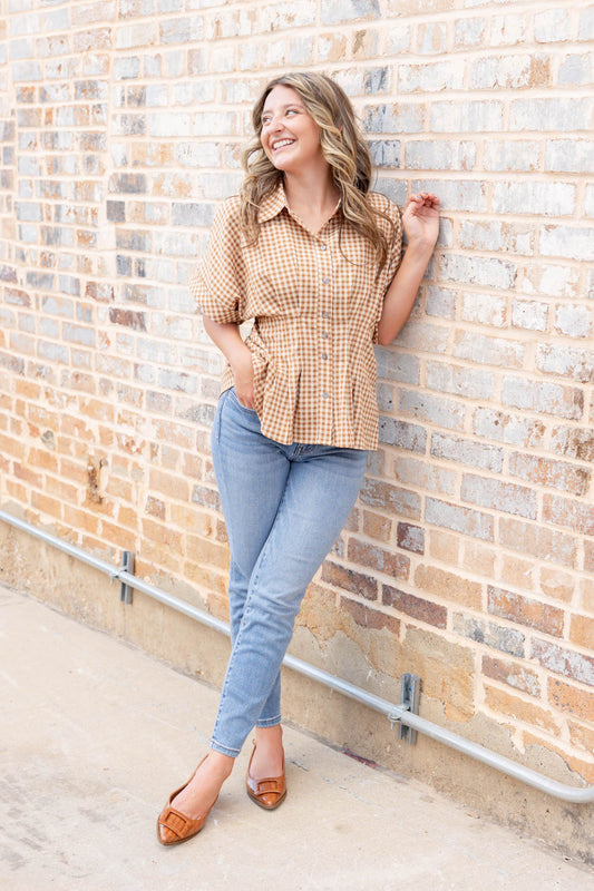 A smiling woman with wavy light brown hair stands against a brick wall, wearing a tailored tan and white gingham short-sleeve button-up shirt, blue skinny jeans, and pointed toe slingback flats.