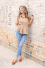 A smiling woman with wavy light brown hair stands against a brick wall, wearing a tailored tan and white gingham short-sleeve button-up shirt, blue skinny jeans, and pointed toe slingback flats.