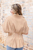 A woman wearing a tan and white gingham short-sleeve blouse with a peplum waist, shown from the back against a brick wall.