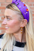 A blonde woman in profile wearing a purple fabric headband adorned with small Halloween-themed charms and white pearl-like beads, paired with dangling orange and white earrings, set against a brick wall background.