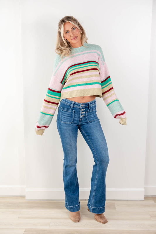 A woman modeling a pastel striped cropped knit sweater and blue jeans in a bright indoor setting with a white wall and light wood floor.