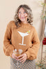 A woman wearing a chunky knit camel brown sweater with a white knitted martini glass graphic on the front, holding a cocktail glass in a warm indoor setting.