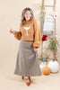 A woman stands in a styled studio setting wearing a camel-colored knit sweater with a white martini glass graphic and a black-and-beige plaid midi skirt. She holds a cocktail glass and is posed with pumpkins, a plaid throw, and floral decor against a warm beige backdrop.