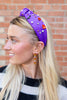A blonde woman in profile wearing a purple headband embellished with pearls, small black beads, and red and orange halloween charms, set against a brick wall background.