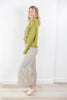 A woman modeling a light olive green knit cardigan and beige-and-blue vertically striped wide-leg trousers, standing in a bright room with white walls and light wood flooring.