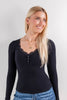A blonde woman wearing a black long-sleeve henley top with a lace-trimmed scoop neck and button details, paired with blue jeans, photographed against a light gray studio background.