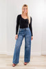 A woman modeling a pair of high-waisted, wide-leg blue jeans with a faded leopard print, paired with a black long-sleeve top and red heel sandals, standing against a white wall and light wood floor.