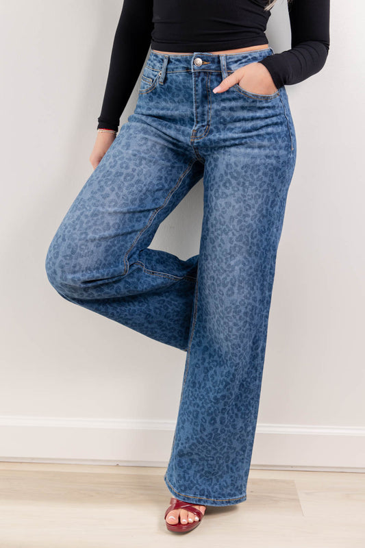 A woman wearing blue denim leopard print wide-leg jeans, with a black long-sleeve crop top, and burgundy open toe heels, standing against a wall and wood floor.