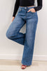 A woman wearing blue denim leopard print wide-leg jeans, with a black long-sleeve crop top, and burgundy open toe heels, standing against a wall and wood floor.