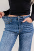 Close-up of a woman wearing blue leopard-print jeans, stretching the waistband to show how loose or tight the fit is, with a black ribbed long-sleeve top against a light studio background.
