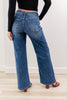 A woman shown from the back wearing blue wide-leg jeans with a subtle tonal leopard print pattern, paired with a black long-sleeve top and red heeled sandals in an indoor room setting.