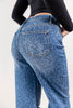Close-up back rear view of a woman wearing blue denim toned leopard-print jeans with a black long-sleeve top, standing against a light gray studio background.