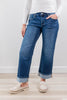 A woman wearing blue straight-leg cuffed jeans with a white long-sleeve top and cream slip-on loafers, standing against a light gray wall and pale wood floor.