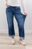 A woman wearing wide-leg blue jeans with thick cuffed hem, paired with a white long-sleeve top and cream slip-on shoes, standing on a light wood floor against a white wall.