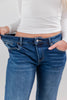 A close-up of a woman wearing low rise blue denim jeans and a white ribbed long-sleeve top, holding the waistband of the jeans wide with both hands to demonstrate the stretchy, baggy, loose fit.