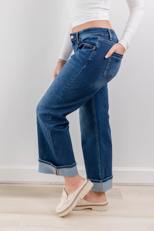 A woman wearing low rise blue cuffed jeans with a white long-sleeve crop top and off-white slip-on loafers, standing in a bright indoor room with a white wall and light wood floor.