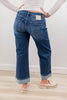 A woman shown from the back wearing wide leg blue denim jeans with rolled cuffs, a white long-sleeve top, and white slip-on shoes in an indoor room with a light wall and wood floor.
