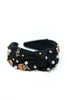 A black fabric headband with a knotted bow design, decorated with small white pearls, gold studs, and colorful enamel halloween pins, shown on a plain white background.