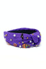 A purple fabric twist-front headband decorated with small white faux pearls, gold-tone studs, a black bat charm, and colorful Halloween-themed enamel pins including pumpkins and a cat.