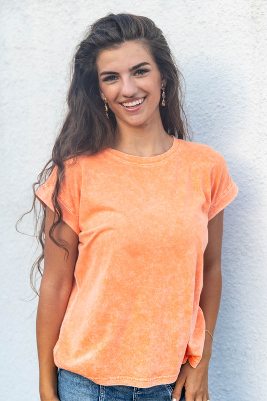 A smiling woman, wearing a short-sleeve crewneck t-shirt in a washed orange color, paired with blue jeans, standing against a white textured wall.