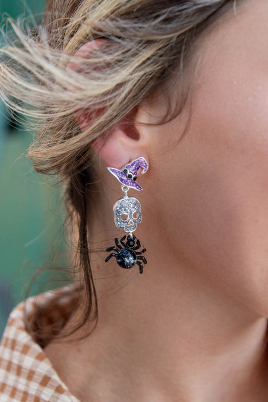 A close-up of a person's ear wearing a dangling Halloween earring with a purple witch hat, a silver skull, and a black spider charm.