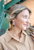 A woman in a brown and white gingham shirt wearing a pair of dangling earrings with a purple crystal heart, a clear crystal ball, and a black spider charm, set against an outdoor background with a green staircase.