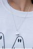 A close-up of a person wearing a delicate silver chain necklace with a small white ghost pendant, styled with a white crewneck sweatshirt featuring a black ghost graphic.
