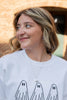 A woman with blonde-highlighted hair wearing a white crewneck sweatshirt with a black line art graphic of three ghosts, with a matching enamel ghost necklace photographed outdoors against a warm-toned brick wall.
