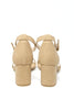 A pair of beige block-heel sandals shown from the back, featuring ankle straps and a small side buckle, set against a plain white background.