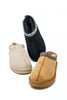 Three suede ankle slipper boots arranged on a white background: one black slipper standing upright in the back with a white patterned trim, one tan slipper in the front with a multicolor patterned trim, and one light beige slipper on the left with a white patterned trim.