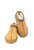 A pair of tan suede slip-on slippers with a soft cream sherpa lining and a decorative patterned trim around the collar, shown on a white background.