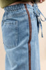 Close-up of a person wearing light blue denim skirt or pants with a vertical brown stripe down the side, an elastic waistband, back pocket, and drawstring tie, against a plain beige background.