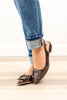 A woman wearing brown pointed-toe flats with large square buckle details and ankle straps, paired with cuffed blue jeans, standing on a light wood floor.