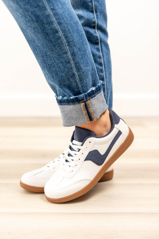 White low-top sneakers with navy blue side stripes and gum soles, shown on a woman wearing blue jeans with the cuffs rolled up, standing on a light wood floor.