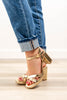 A woman wearing metallic gold strappy block-heel sandals with blue cuffed jeans, standing on a light wood floor against a white wall.