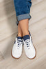 White low-top sneakers with navy blue accents and brown gum soles, shown on a woman's feet wearing cuffed blue jeans against a light wood floor background.